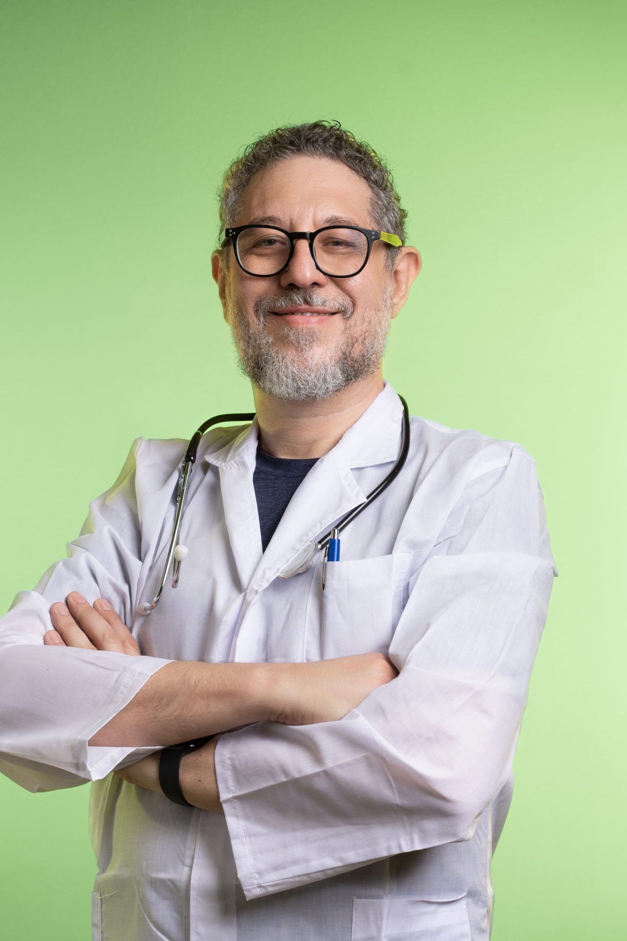 Smiling male doctor with a beard and glasses, wearing a lab coat and stethoscope, standing confidently with arms crossed, light green background, representing trust and professionalism in healthcare
