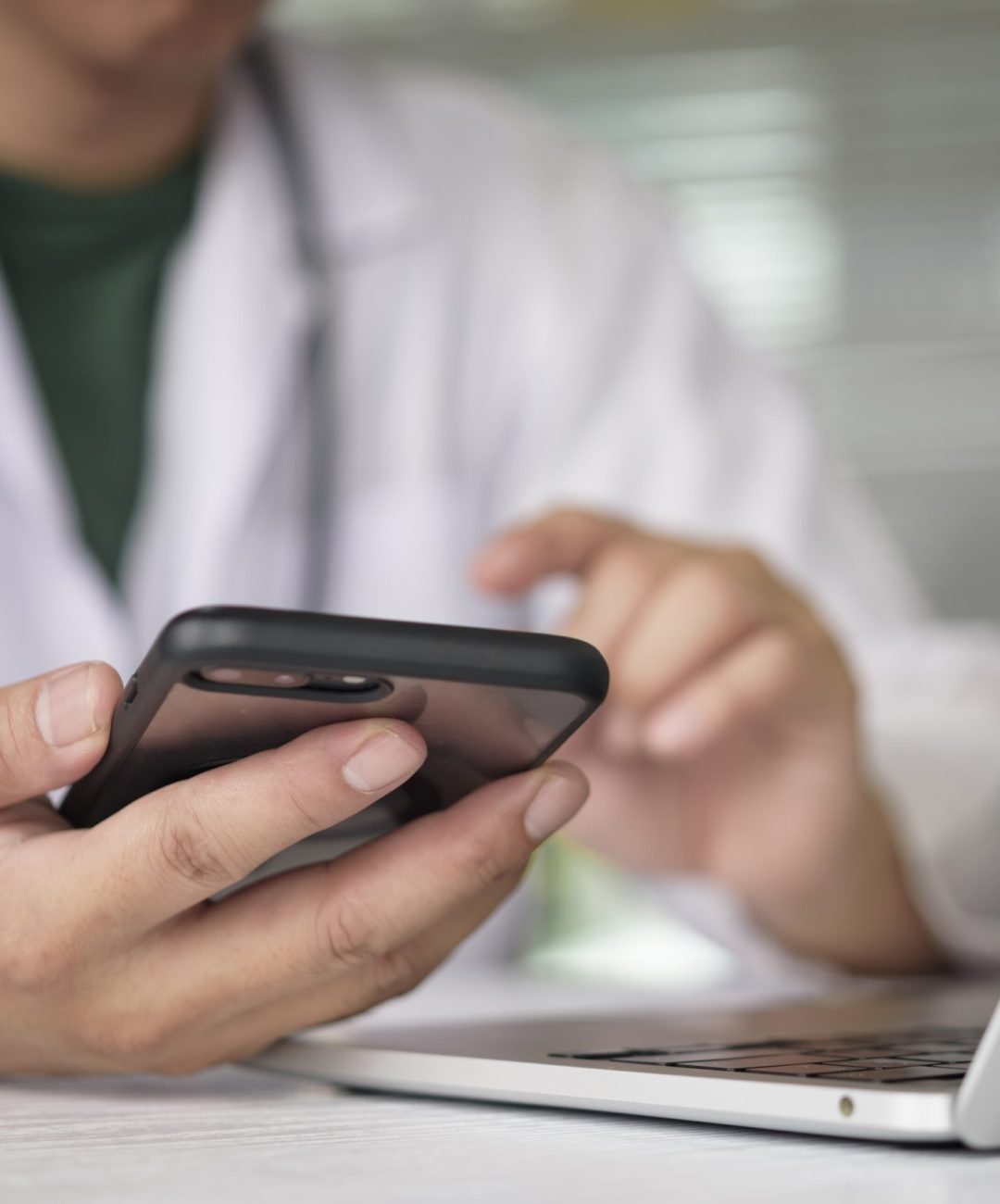 Doctor using smartphone and laptop for online consultation with patient. Physician texting on mobile phone in hospital office. Telemedicine, telehealth and digital healthcare technology concept.